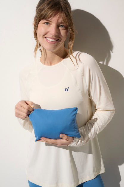 Femme souriante portant une veste anti-UV beige UPF50+, tenant un petit sac bleu, idéale pour activités de plein air.