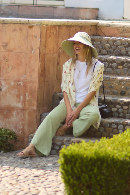 Femme en pantalon anti-UV léger UPF 50+, chapeau et top, assise dans un jardin ensoleillé, idéale pour l'été.