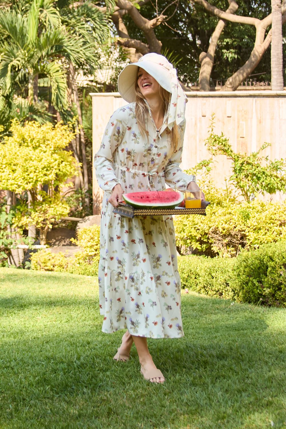 Femme portant une robe longue anti-UV à manches longues, motif floral et chapeau, dans un jardin ensoleillé.